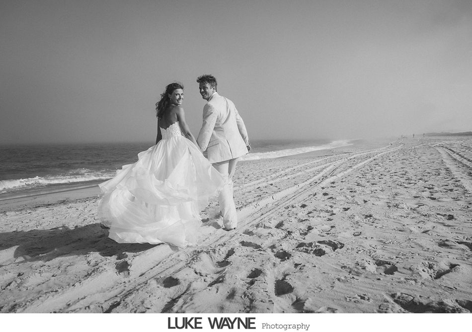 Bride and groom on a beach, holding hands, looking back at the camera. Black and white photo.