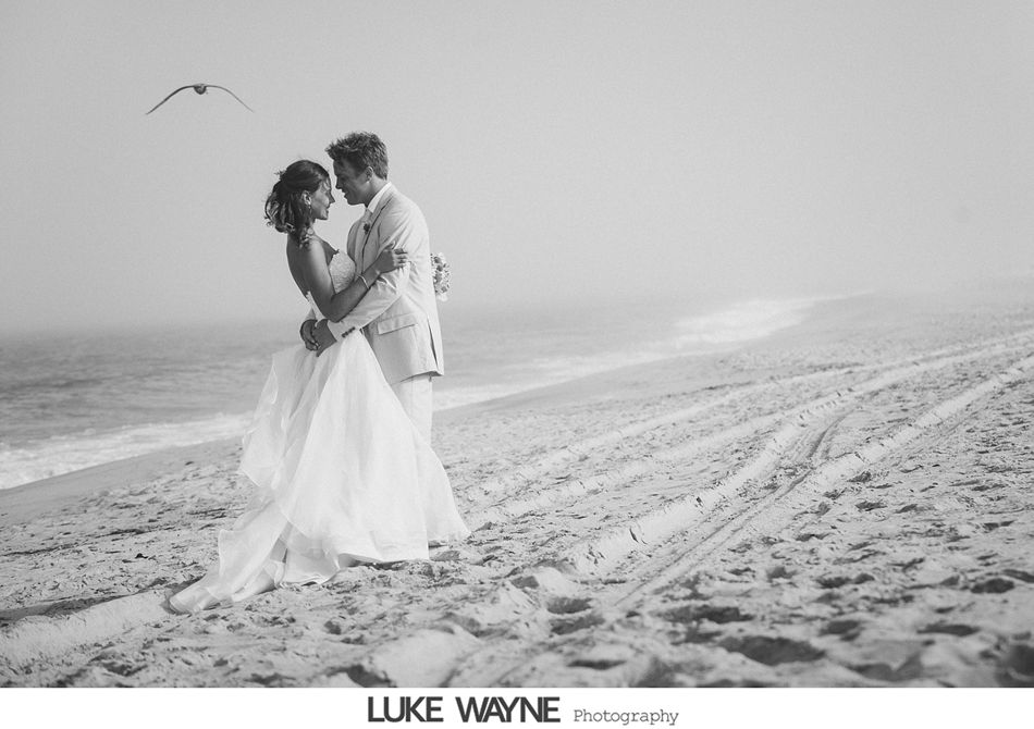 Bride and groom embrace on a misty beach. Seagull flies overhead. Black and white.
