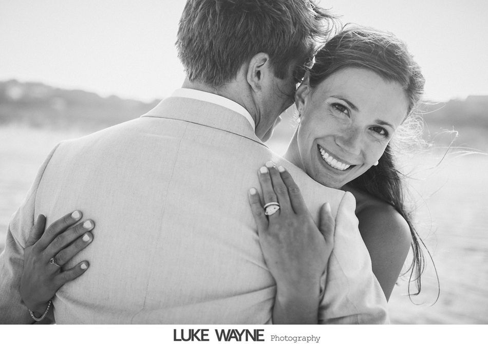 Bride hugs groom, smiling. Bride's engagement ring visible. Beach setting, black and white.