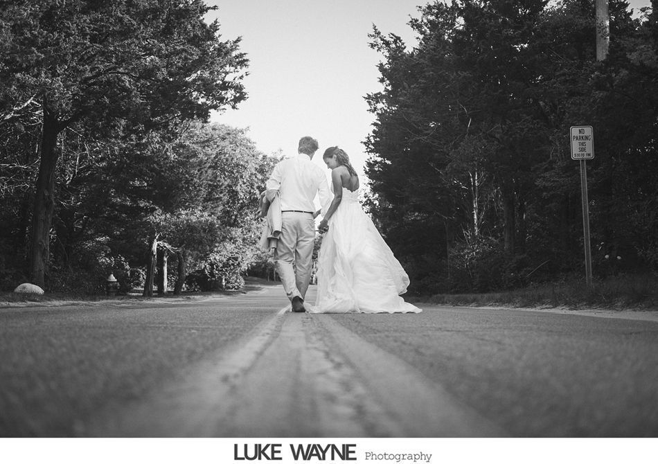 Newlyweds walk down a tree-lined road. The bride wears a gown, the groom a button-down shirt. Black and white.