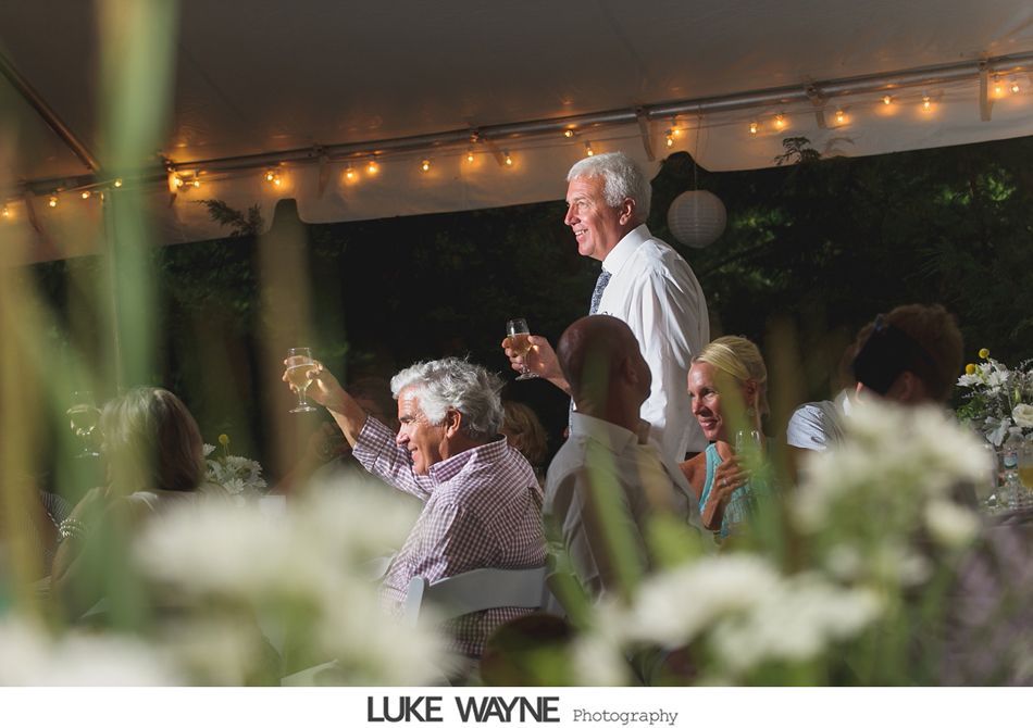 Guests raise glasses at an outdoor wedding reception. Man in white shirt gives a toast under tent with string lights.