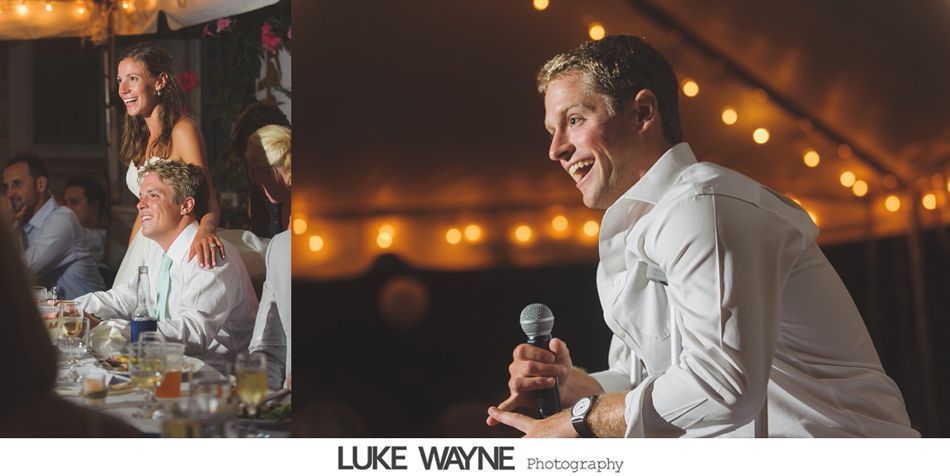 Man speaking at a wedding, holding a microphone, smiling under string lights.