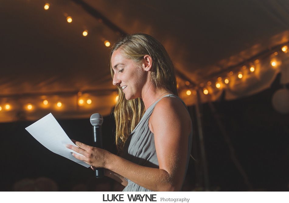 Woman giving a speech, holding paper and microphone, under string lights.