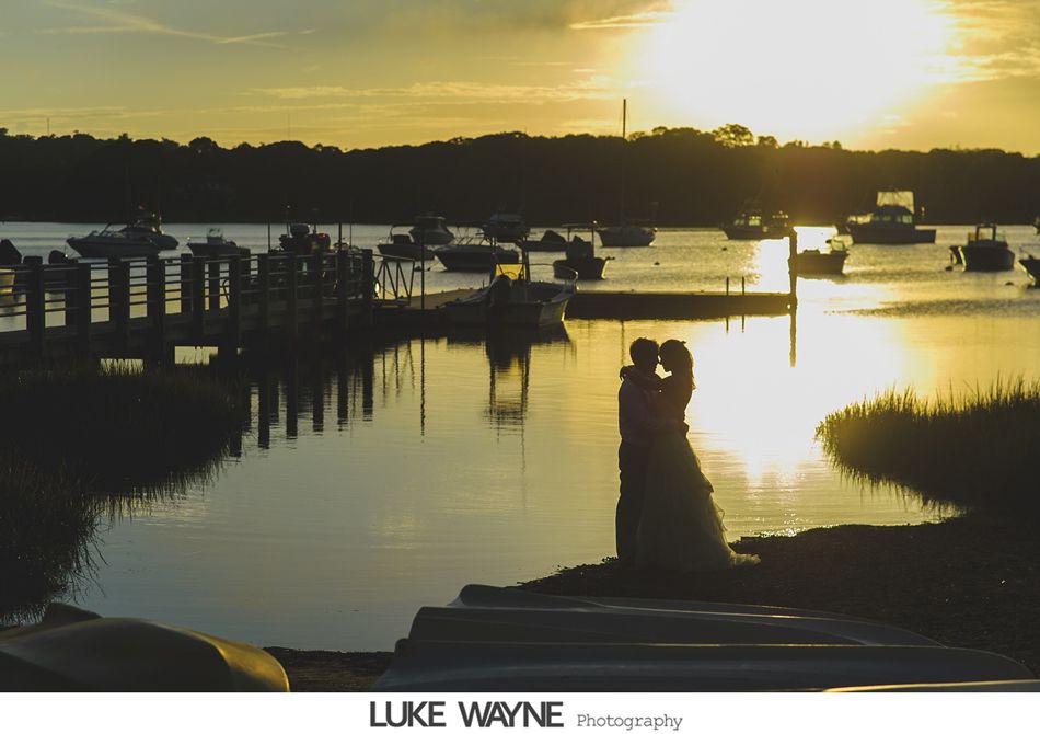 Couple embraces at sunset by a harbor with boats. Silhouetted figures, golden light.