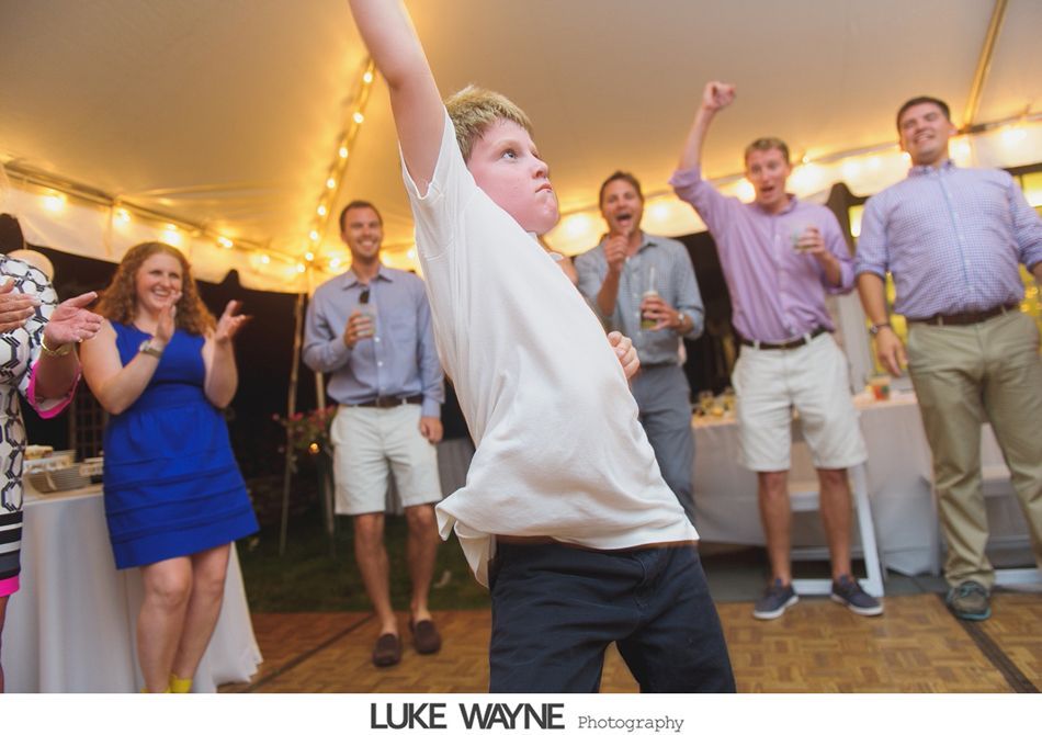 Boy dancing energetically, arm raised, surrounded by cheering people under a tent with string lights.