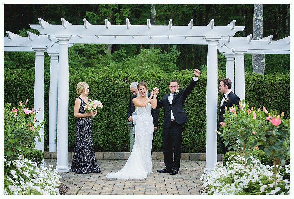 Bride with arms up, making a funny face, groom looking at his hand, cake cutting in background.
