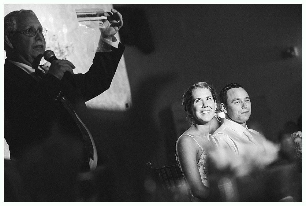 Bride with arms up, making a funny face, groom looking at his hand, cake cutting in background.