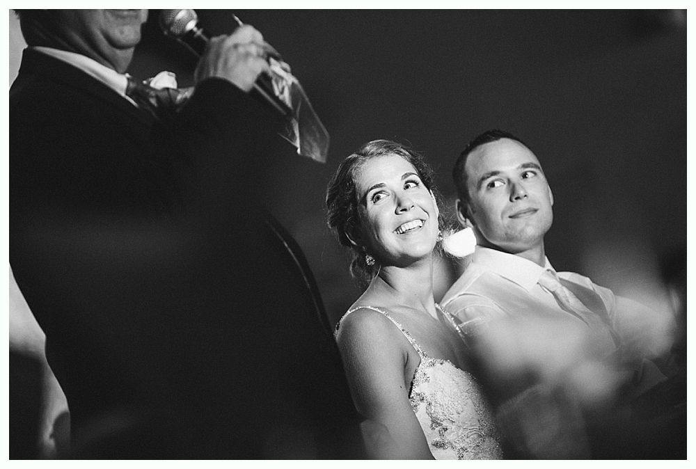 Bride with arms up, making a funny face, groom looking at his hand, cake cutting in background.