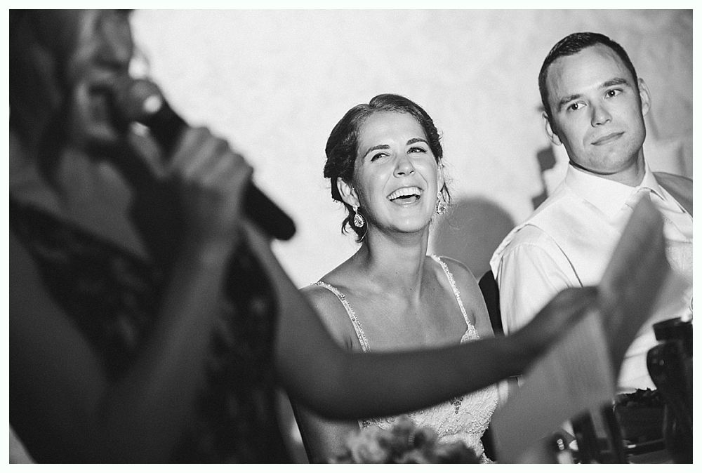 Bride with arms up, making a funny face, groom looking at his hand, cake cutting in background.