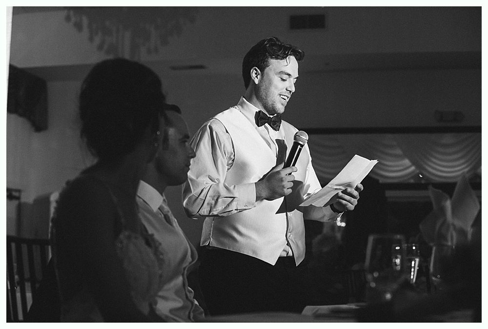 Bride with arms up, making a funny face, groom looking at his hand, cake cutting in background.