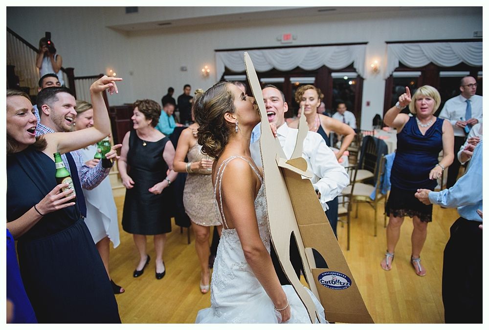Bride with arms up, making a funny face, groom looking at his hand, cake cutting in background.