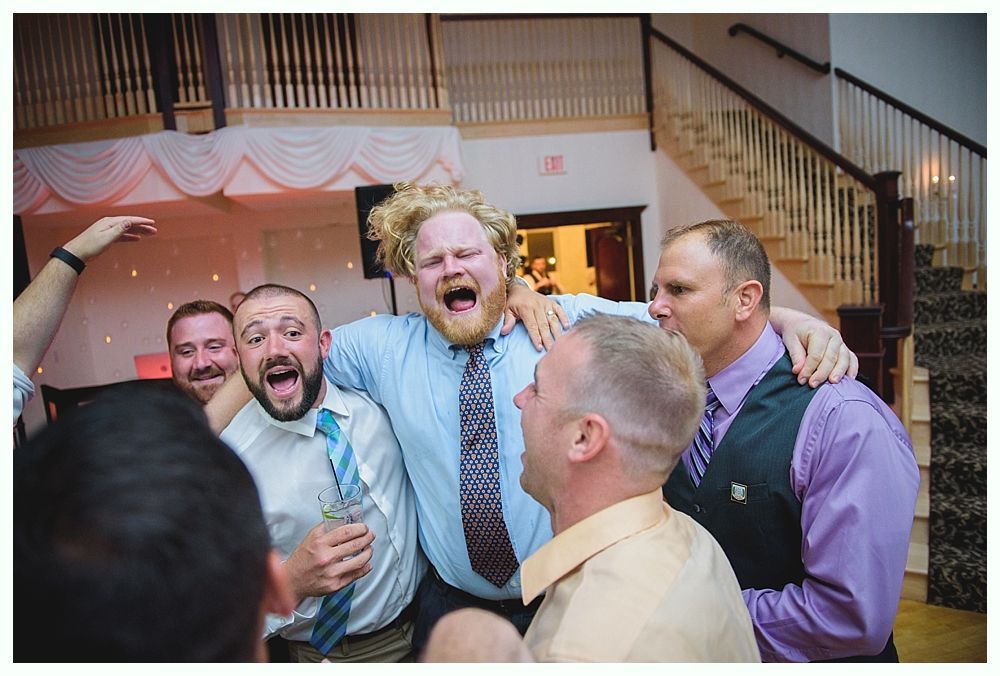 Bride with arms up, making a funny face, groom looking at his hand, cake cutting in background.