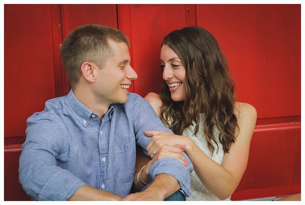 Bride and groom embrace on rocks by the ocean at sunset. The groom wears a blue suit, the bride a white dress.