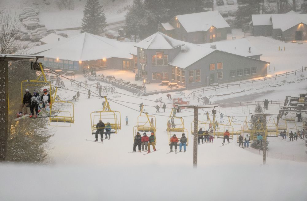 Snowy ski slope with skiers and a chairlift. Buildings and cars in the background.