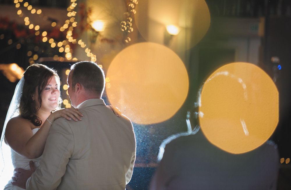 Bride and groom walk down sidewalk, groom smiles, bride holds purple bouquet, city background.