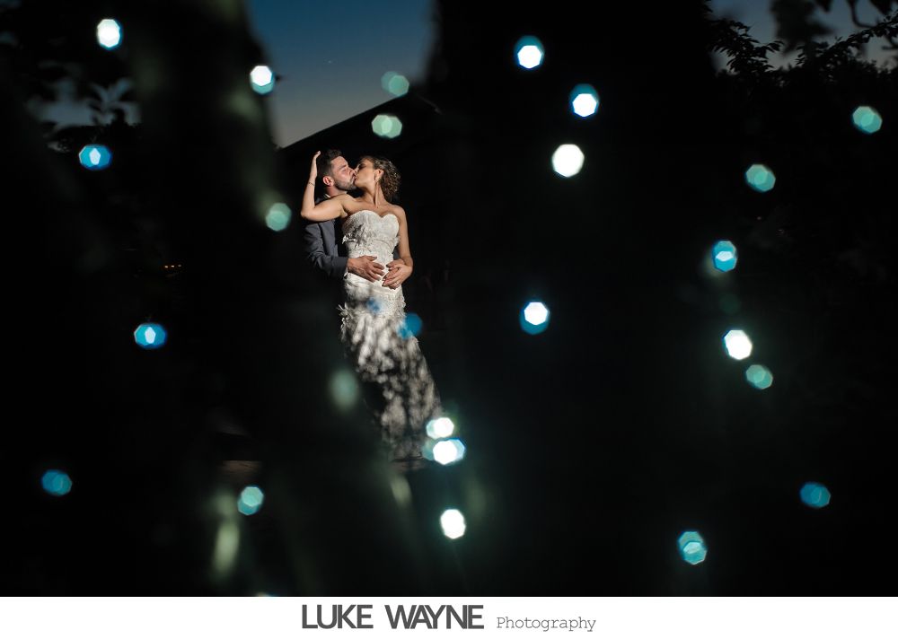 Bride and groom pose near dart boards. Bride in white dress, groom in suit. Indoor setting, red velvet ropes.