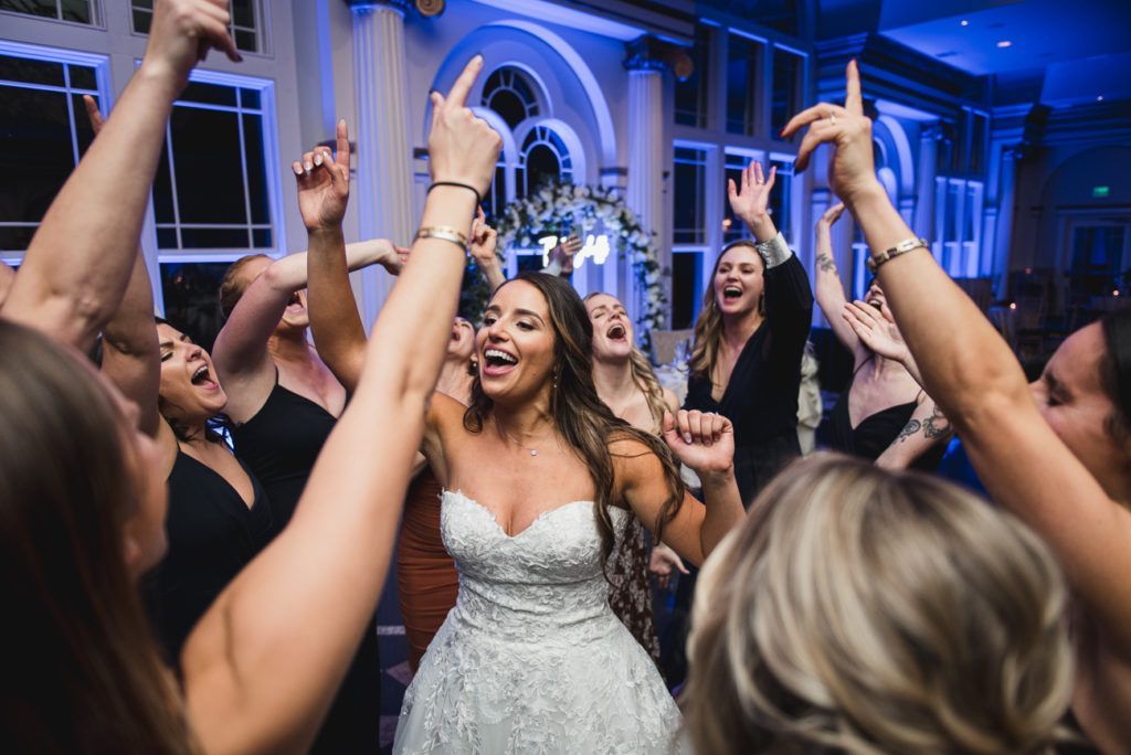 Bride dancing with friends at a wedding reception, all raising arms and smiling in a room with blue lighting.