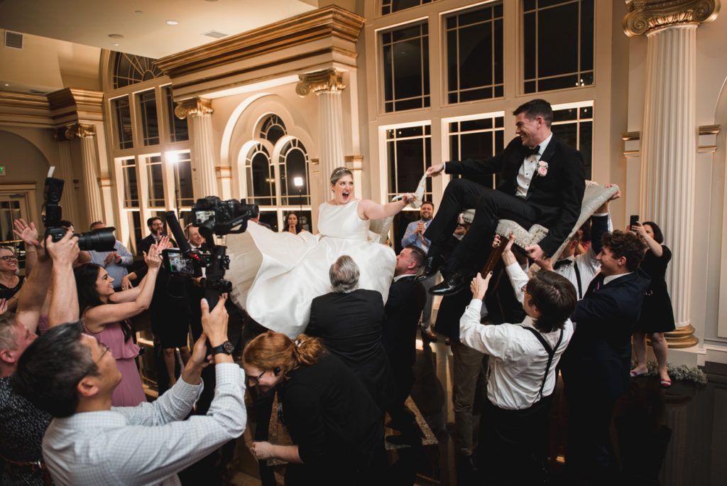 Bride and groom lifted on chairs by guests during wedding reception in a ballroom with ornate architecture.