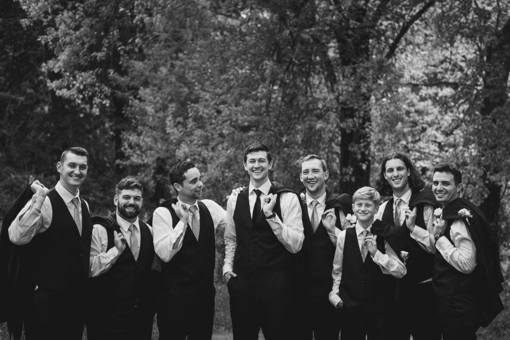 Group of men in vests, smiling and gesturing, outdoors in front of trees.