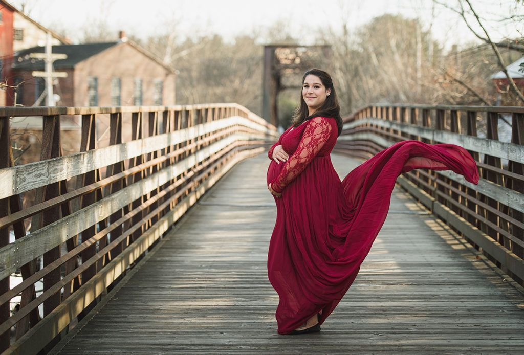 Pregnant person in a flowing red dress stands on a wooden bridge, holding stomach; dress train blowing in the wind.