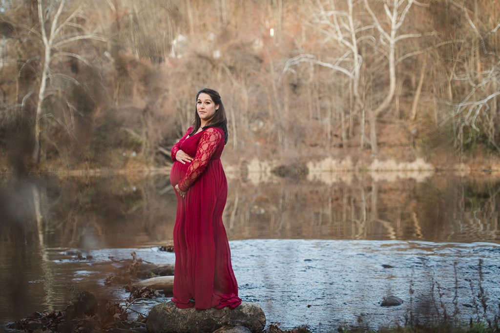 Pregnant woman in red dress stands on rock near water, trees reflected in background.
