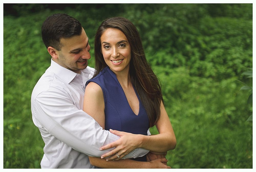 Photographer giving thumbs up to bride adjusting veil, with man in suit and wedding party in background.