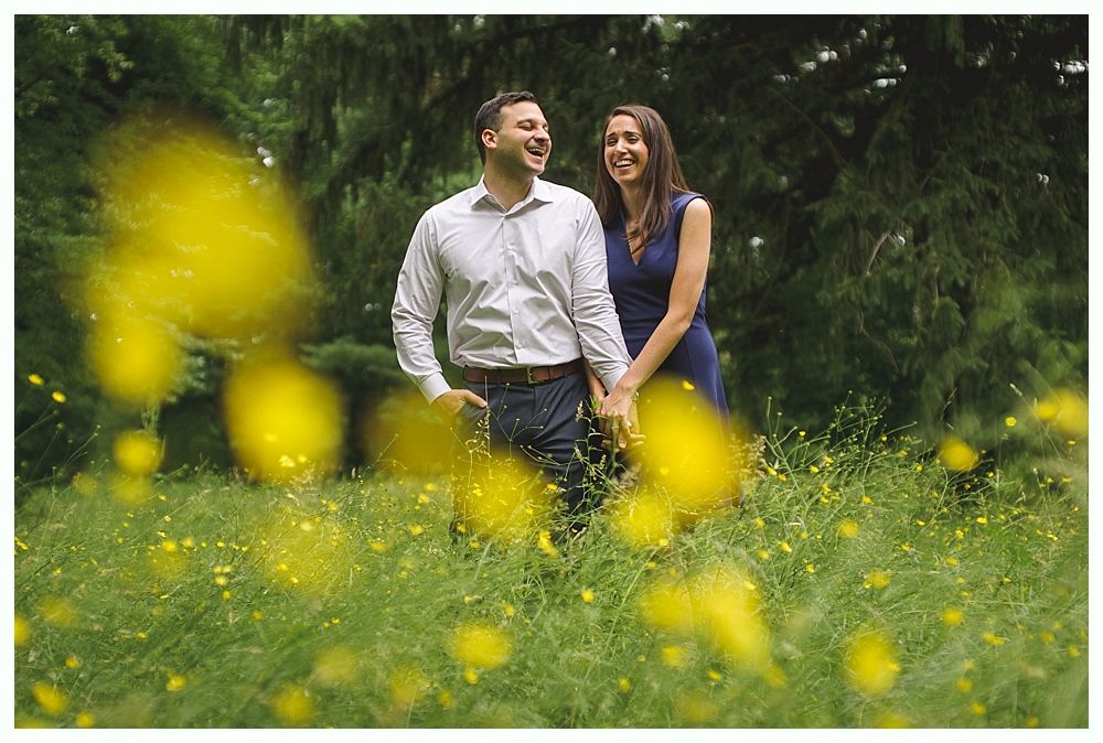 Groom smiles at bride in white dress on bridge, bridesmaids and flowers nearby.