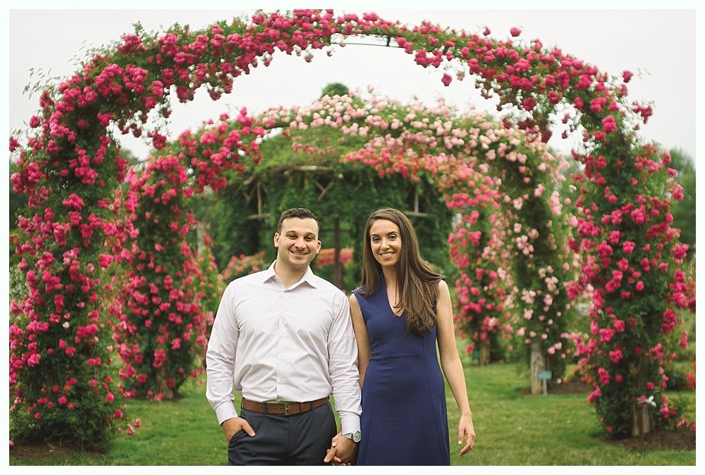 Two women in pink dresses laughing on a bridge with flowers; wedding party in the background.