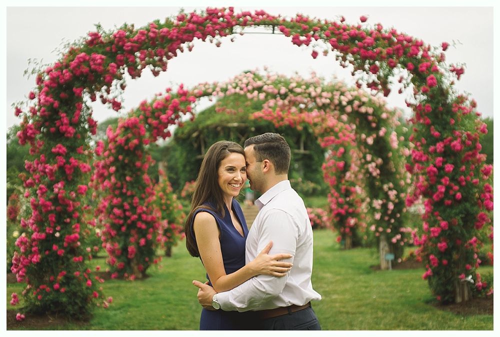 Bride smiles at groom, holding a bouquet. She wears a white dress and veil. He wears a blue suit, looking at her.