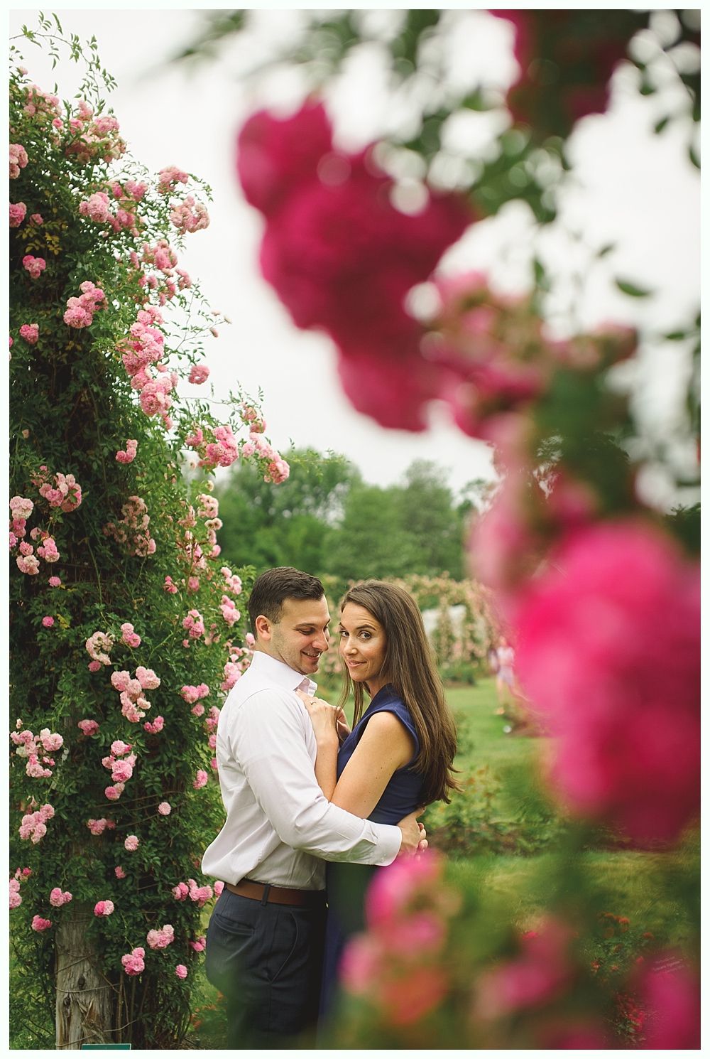 Bride and groom in embrace; the bride gazes at the camera while the groom holds her. Soft background of greenery.