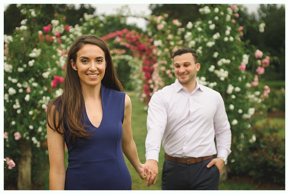 Groom and groomsmen on bridge, wearing suits and pink ties. Groom smiling, holding jacket. Building in background.