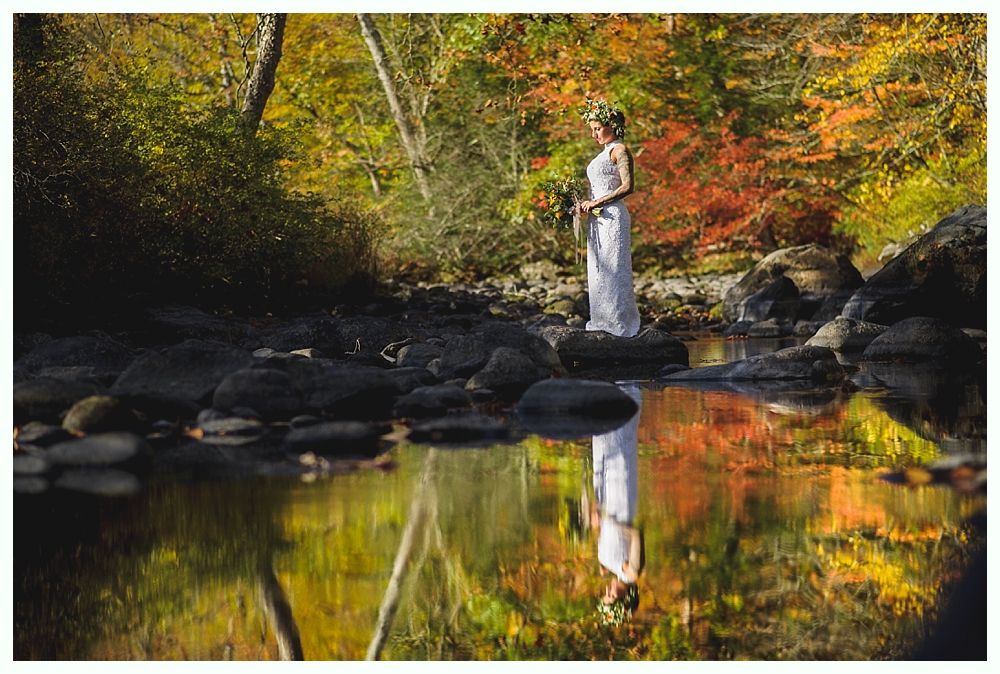 Woman in white dress stands on a rock in a stream, reflected in the water; surrounded by colorful autumn trees.