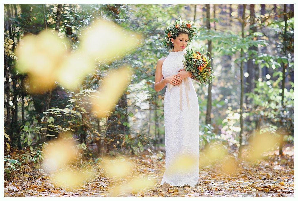Woman in white dress with flower crown holding bouquet in a forest with falling leaves.