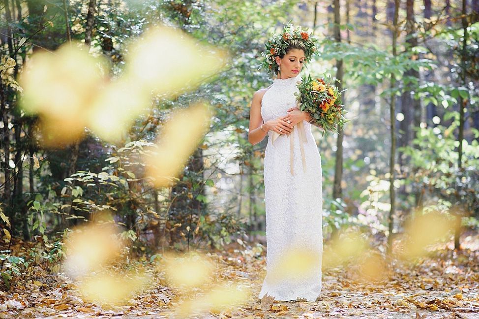 Woman in white dress and flower crown holding bouquet in a forest with out-of-focus yellow leaves.