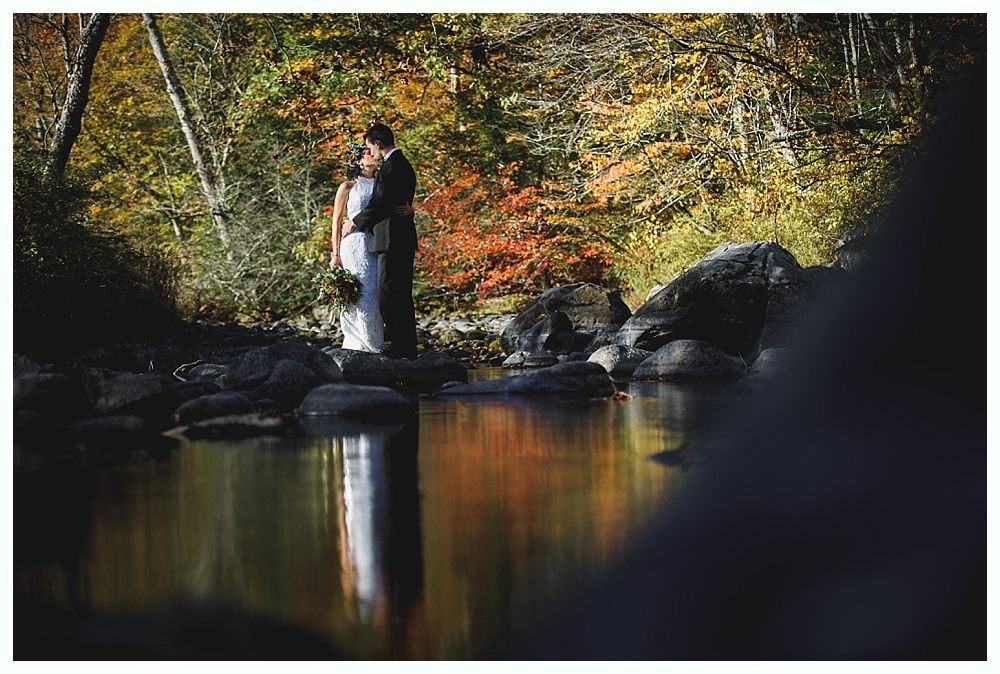 Couple embraces on rocks, reflected in calm river, surrounded by fall foliage.