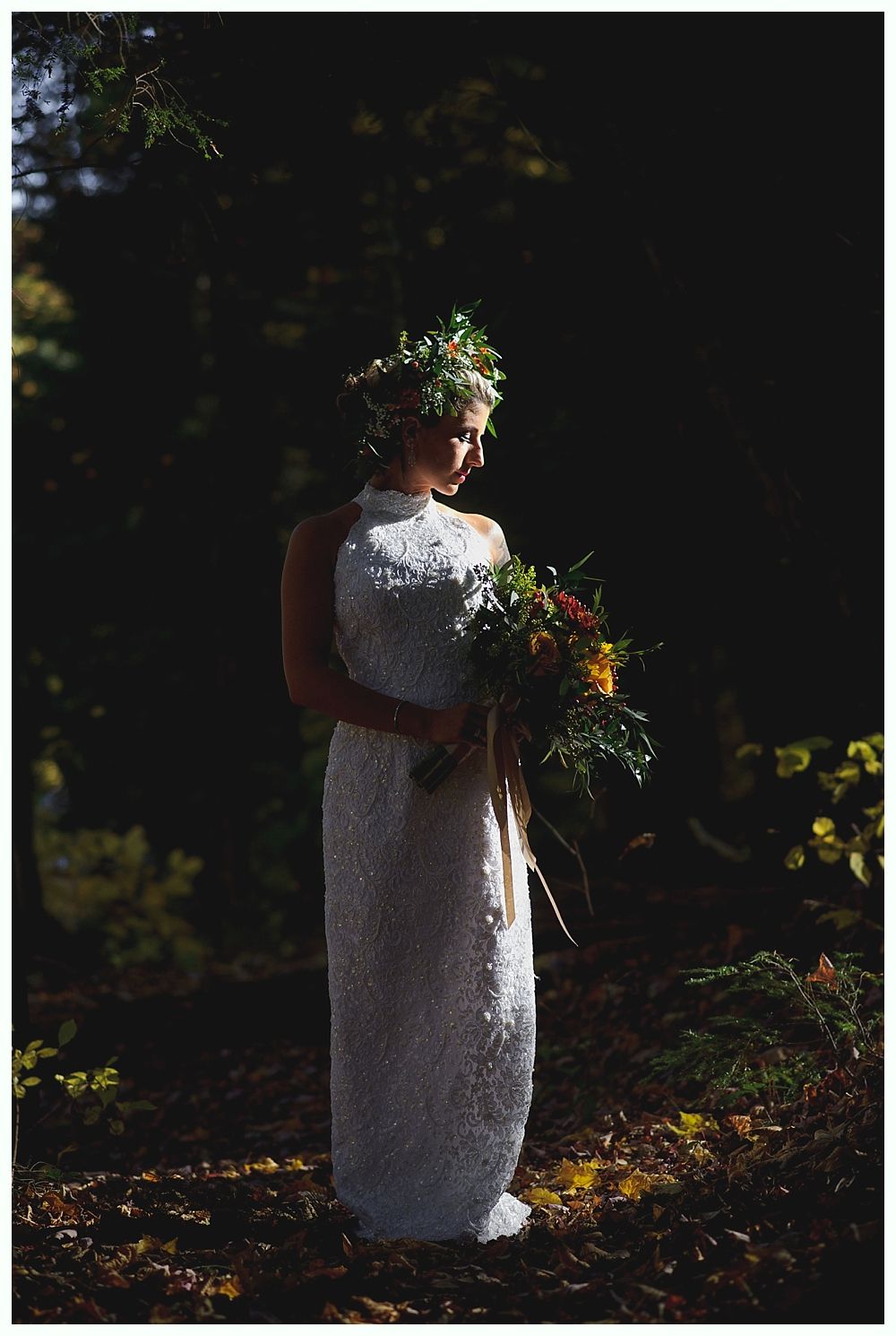 Bride in white beaded dress and floral crown, holding bouquet, in a sunlit forest.