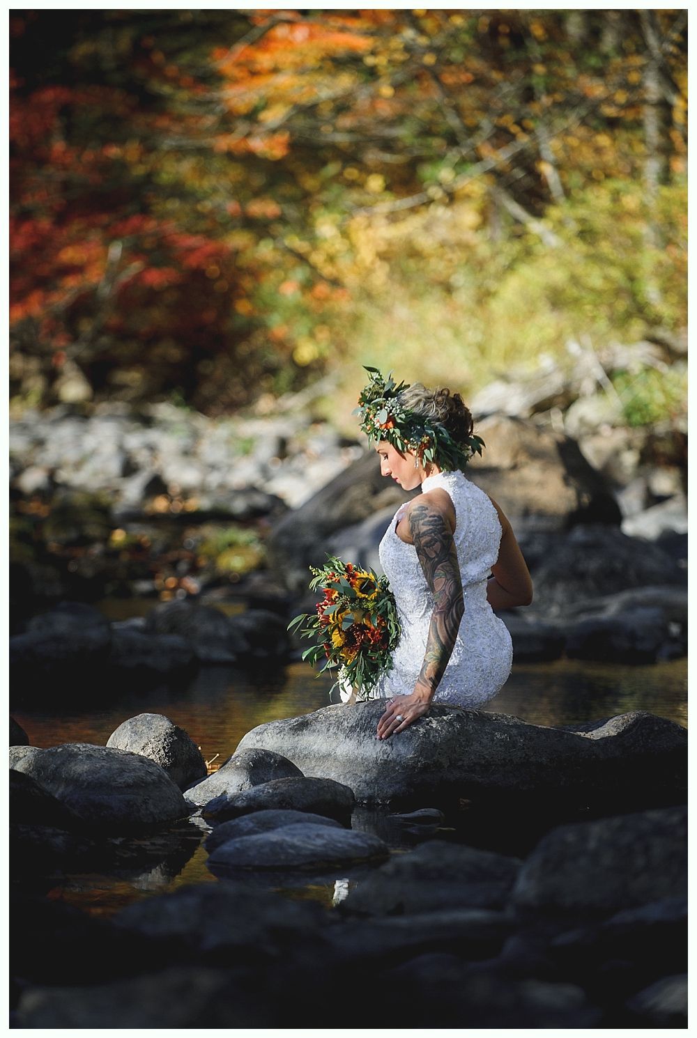 Woman in white dress with flower crown sits on a rock holding a bouquet in a stream, surrounded by fall foliage.