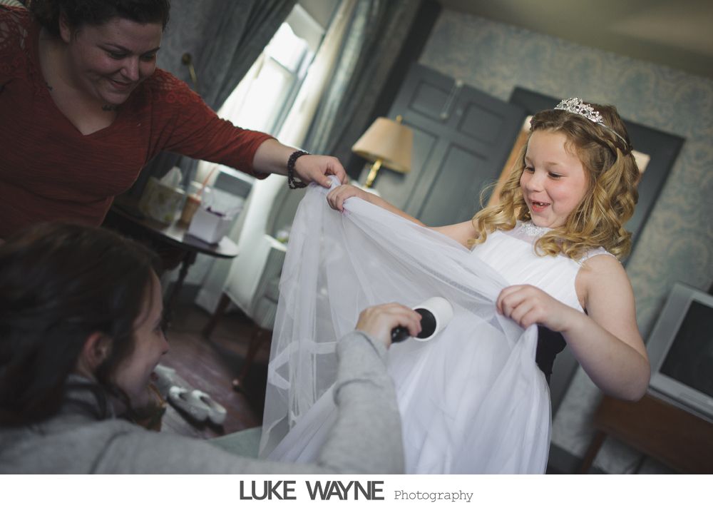 Girl with tiara and white dress smiles as it's being brushed while a woman holds it and another person brushes it; room setting.