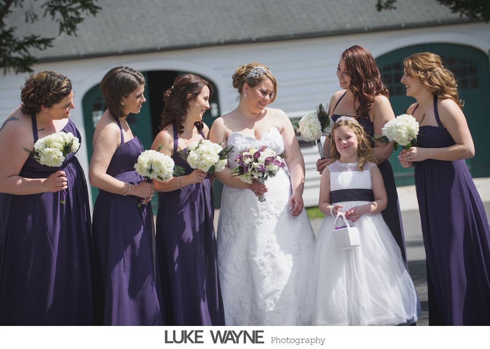 Bride surrounded by bridesmaids in purple dresses, flower girl, holding bouquets, outside a white building.