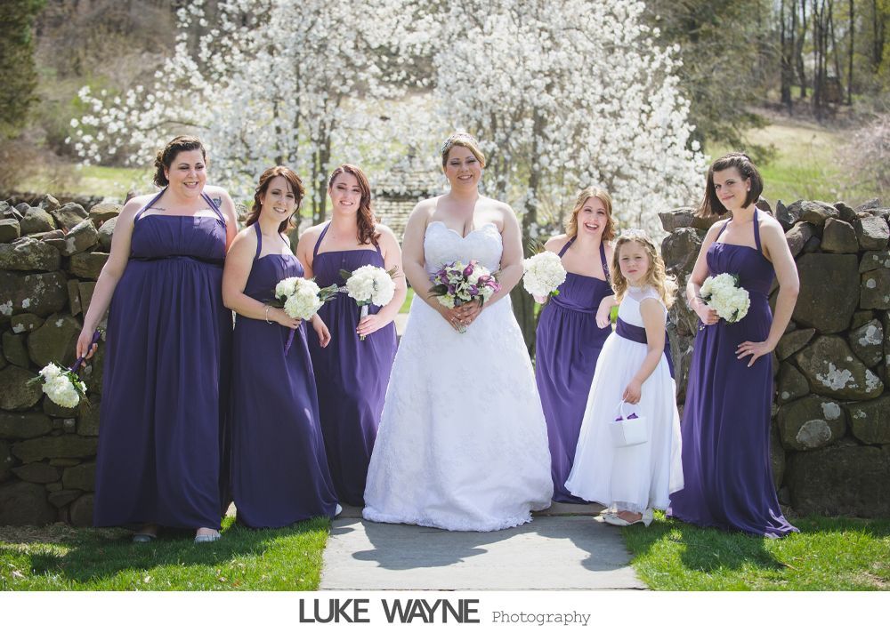 Bride and bridesmaids in purple dresses pose outside, stone wall, white flowers in the background.