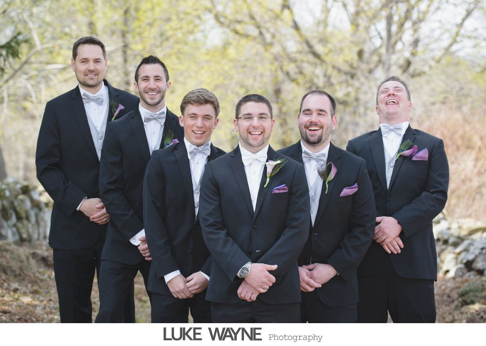 Groomsmen in black suits and bow ties smile for the camera outdoors, with a natural background.