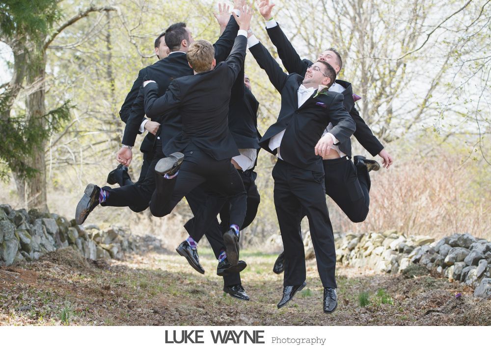 Men in suits jumping and high-fiving outdoors near a stone wall and trees.