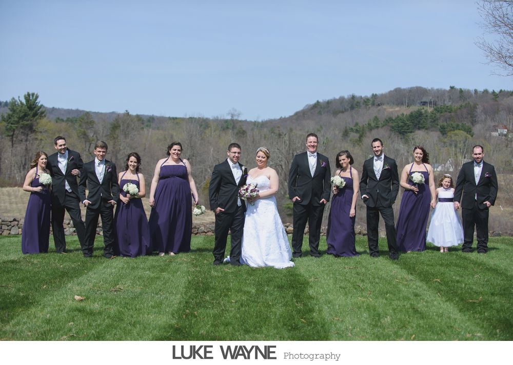 Wedding party stands on grass in front of a hillside. Bridesmaids wear purple dresses, groomsmen wear black tuxedos.
