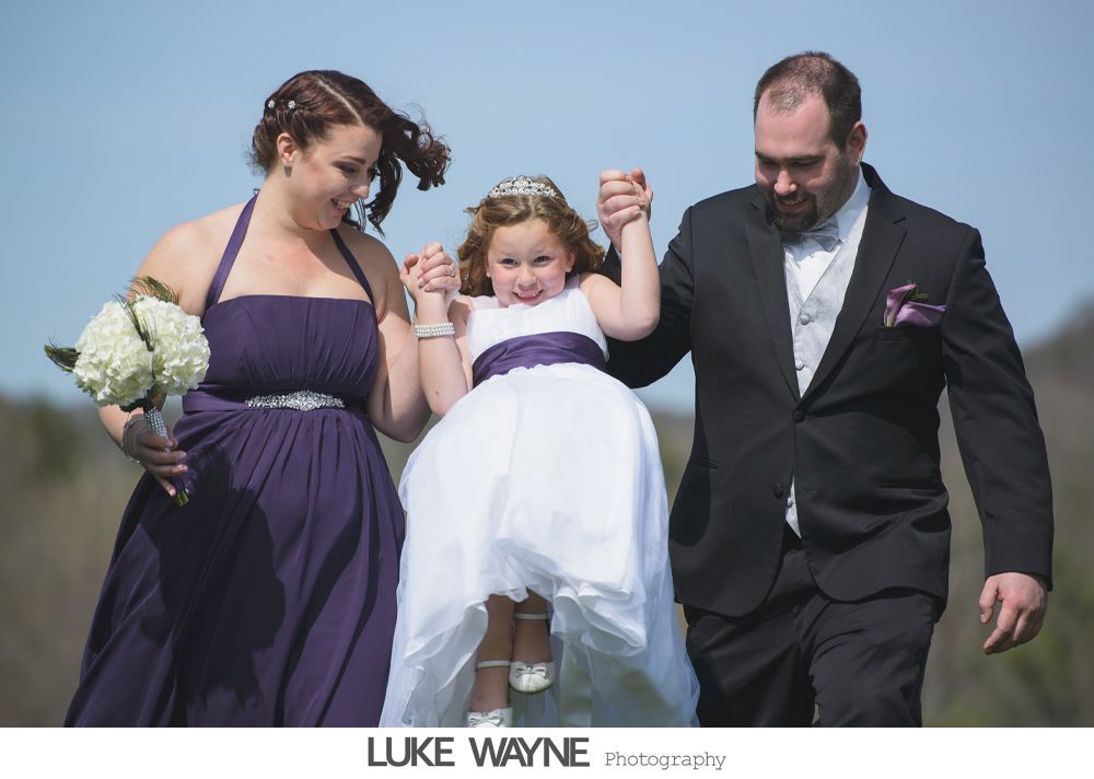 Woman in purple dress and man in suit flank girl in white dress, holding hands, walking outside.