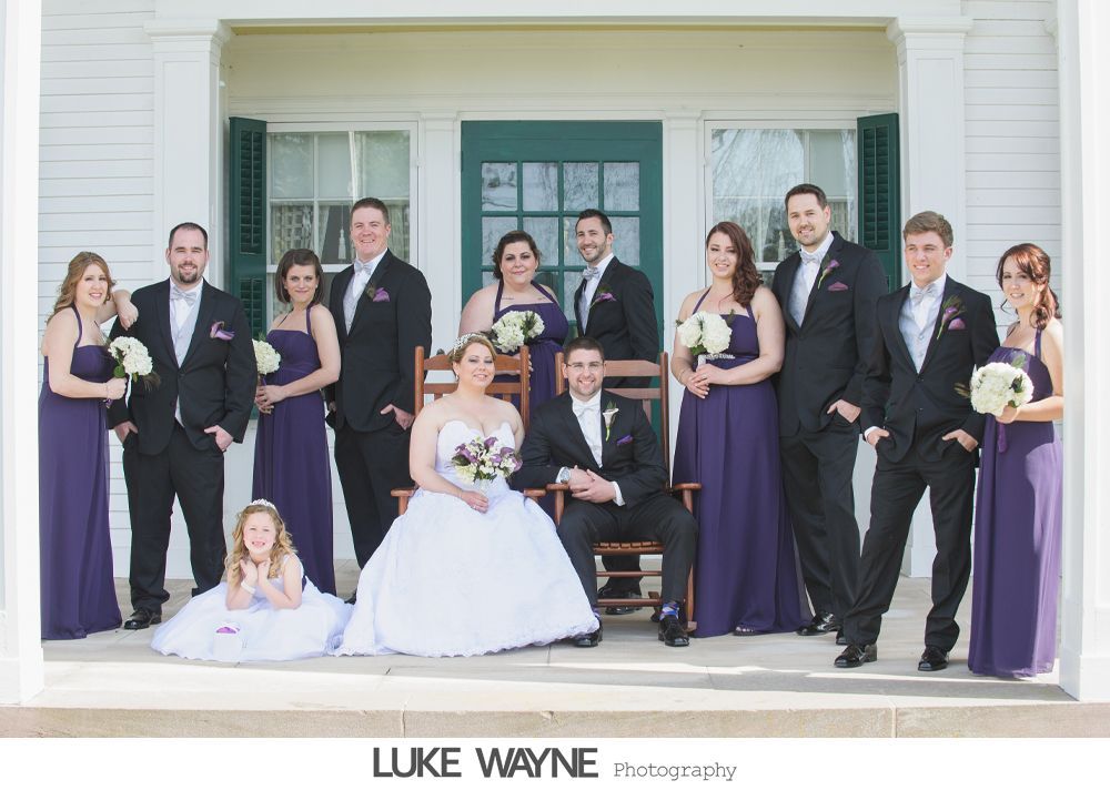 Wedding party posing on porch; bride and groom seated. Purple dresses, black tuxedos. White house background.