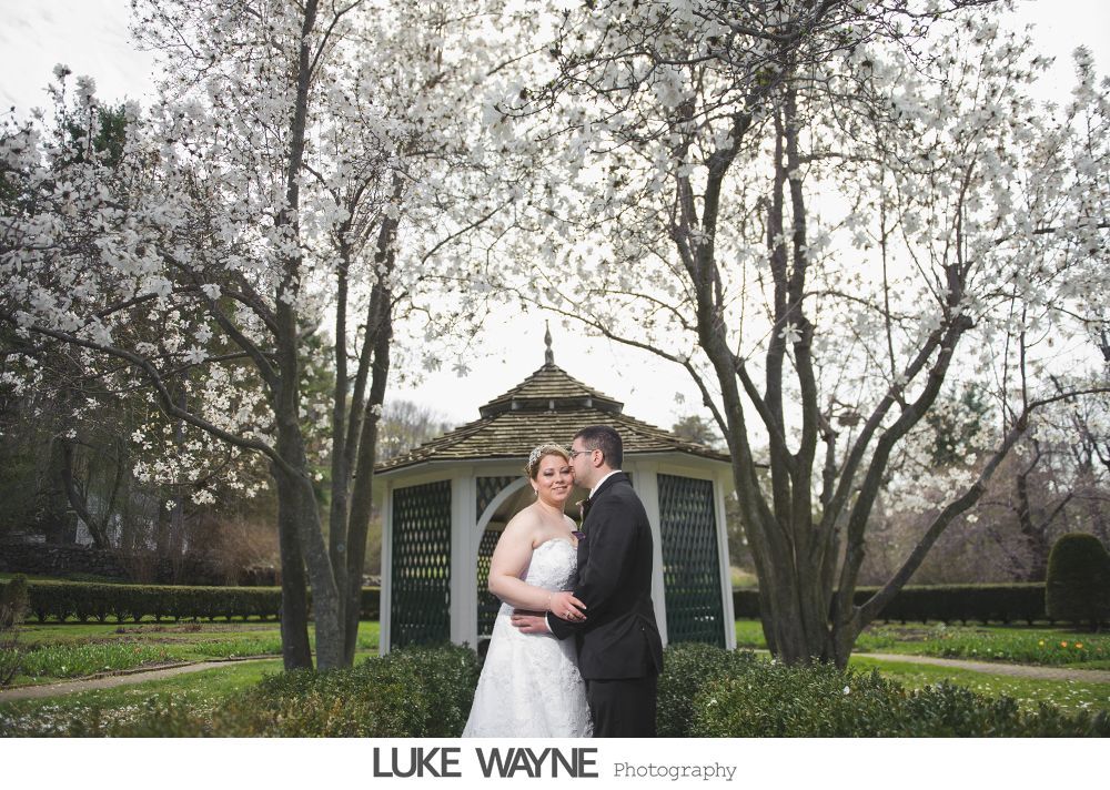 Bride and groom embrace in front of a gazebo, framed by flowering trees.