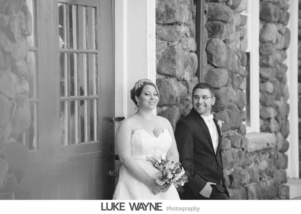 Bride and groom pose by stone building. Bride holds bouquet, groom smiles, both happy.