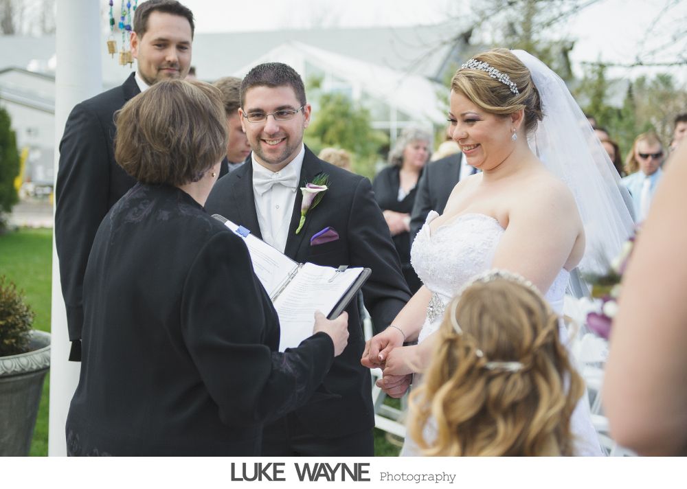 Wedding ceremony: Couple holding hands, smiling at officiant. Guests watch outdoors.