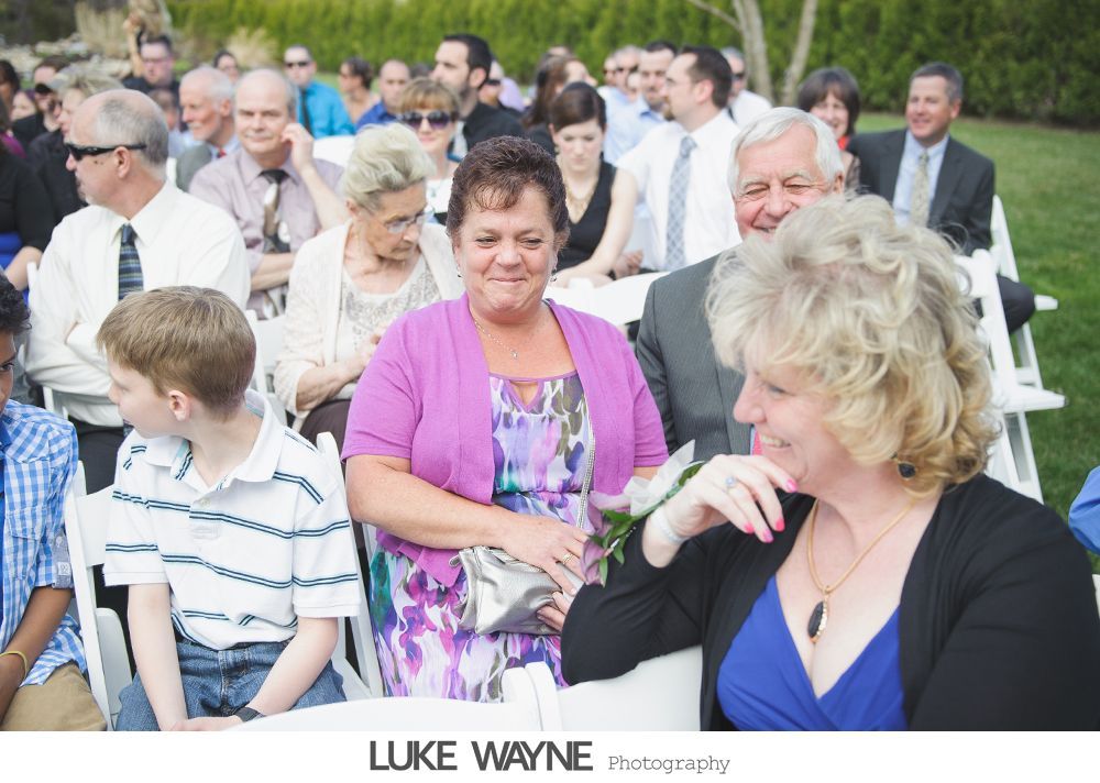 People seated outdoors, possibly at a wedding. Woman in purple smiles, others watch with interest. Green grass and bright day.