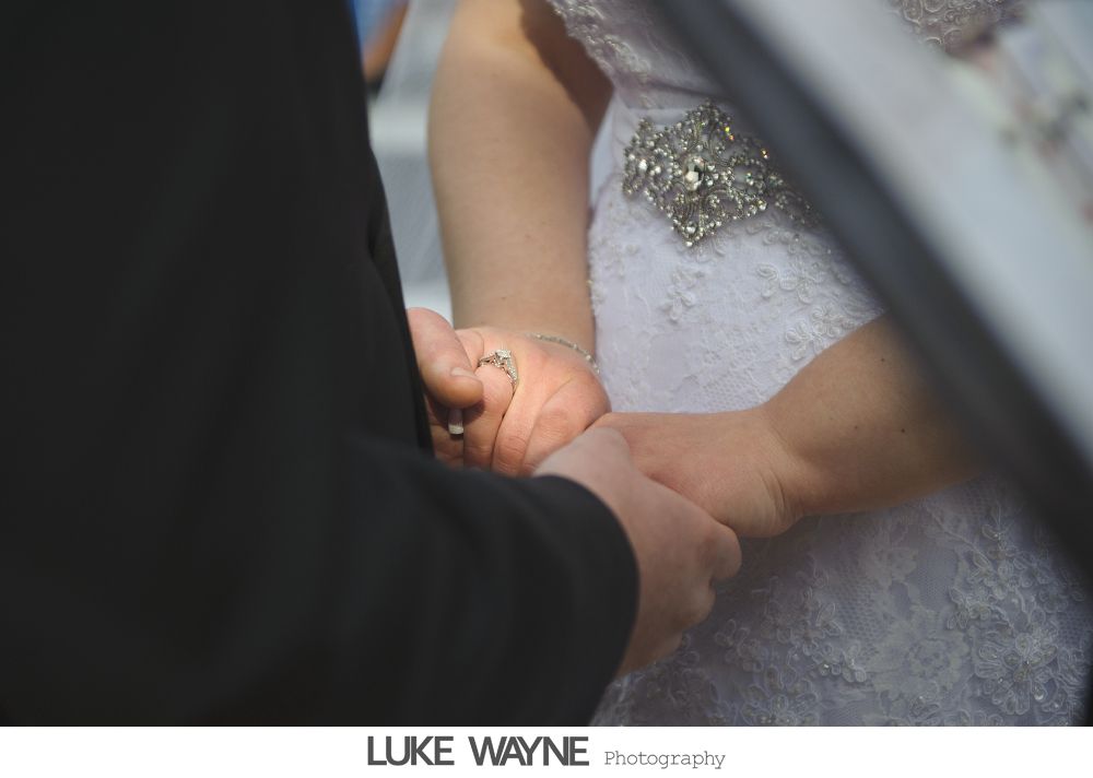Couple holding hands during a wedding ceremony. One is wearing a suit, the other a white dress with a jeweled embellishment.
